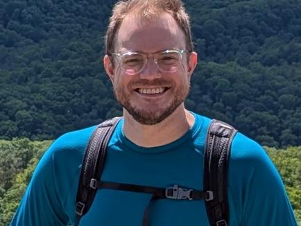 A smiling man with short reddish-brown hair, a beard, and clear-framed glasses poses for a medium close-up outdoors. He is wearing a teal long-sleeved athletic shirt and a black hiking backpack with the chest strap buckled. The background features a vast, sunlit landscape of rolling hills covered in dense green forest under a bright sky.