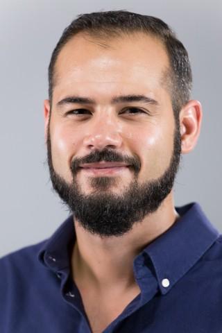 A headshot of a smiling man with short dark hair, a full dark beard and mustache, wearing a dark blue button-up shirt against a plain light gray background.