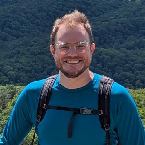 A smiling man with short reddish-brown hair, a beard, and clear-framed glasses poses for a medium close-up outdoors. He is wearing a teal long-sleeved athletic shirt and a black hiking backpack with the chest strap buckled. The background features a vast, sunlit landscape of rolling hills covered in dense green forest under a bright sky.