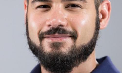 A headshot of a smiling man with short dark hair, a full dark beard and mustache, wearing a dark blue button-up shirt against a plain light gray background.